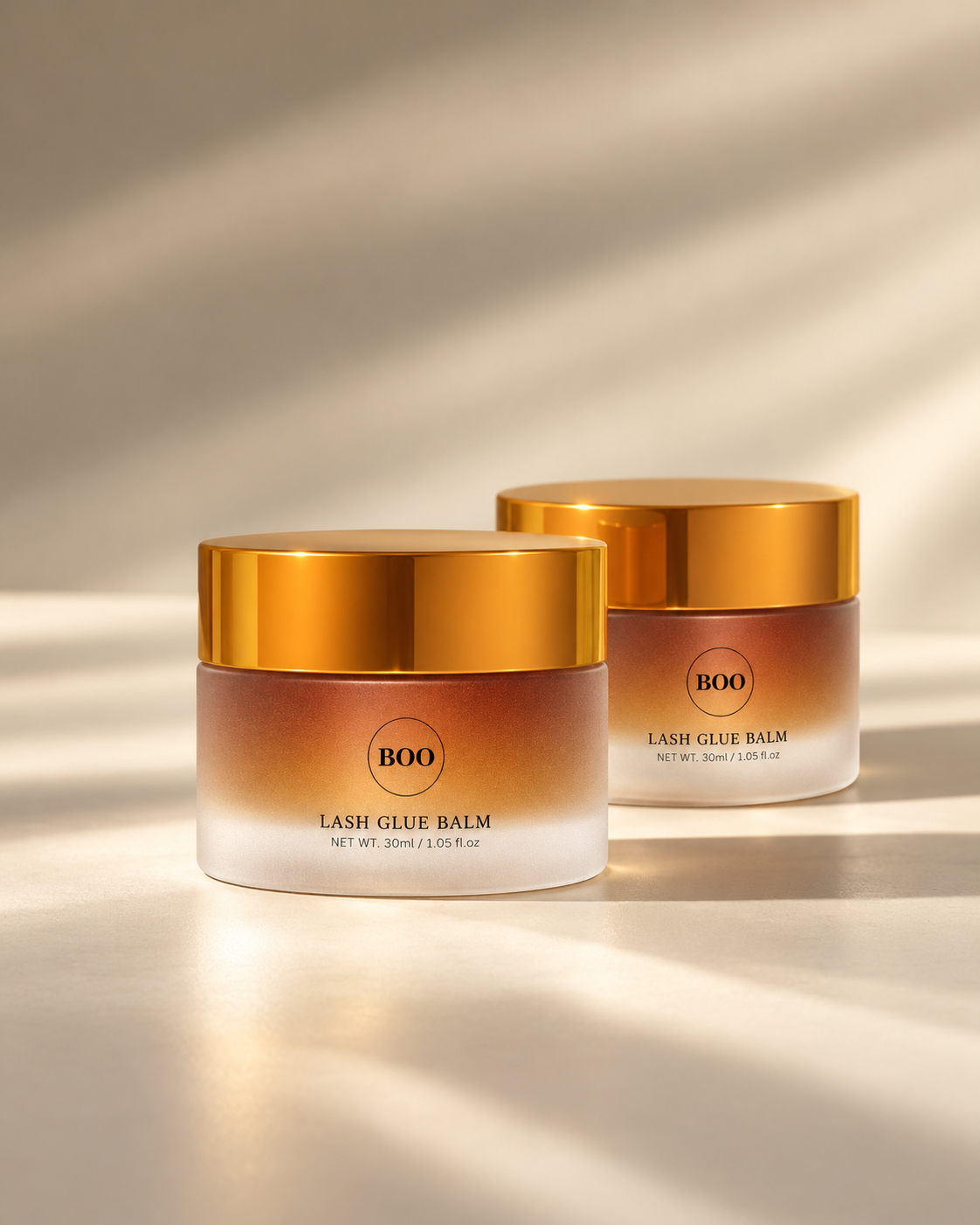 Two closed BOO Lash Glue Balm jars in a luxury gold and ivory still life